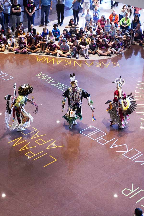 overhead shot of students sitting on the ground and three American Indians dancing in the center in the Bullock Museum